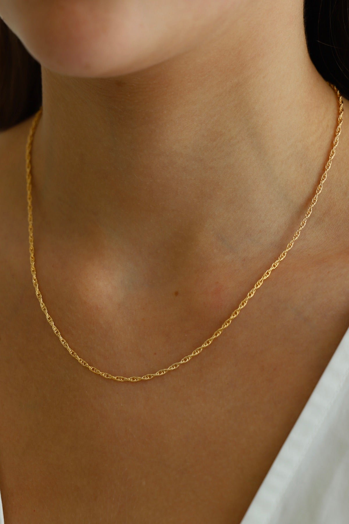 A delicate gold chain necklace rests on a woman's neck, catching the light. The skin appears smooth and toned, contrasting subtly with the shiny jewelry. The background is softly blurred.