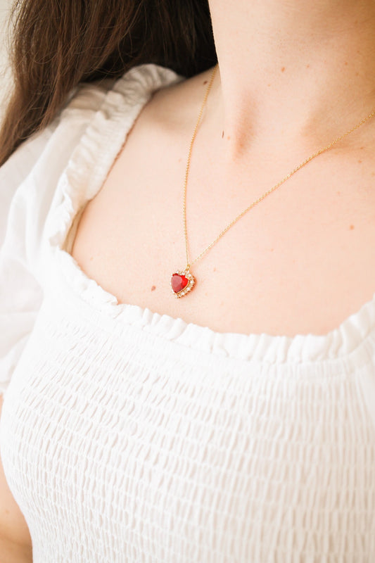 A gold necklace with a heart-shaped red pendant rests gently on a person's collarbone, while they wear a white, textured blouse with a wide neckline.