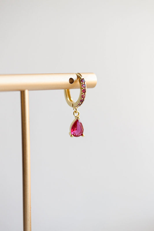 A golden earring with a teardrop-shaped red gem hangs from a display stand. The simple, light background emphasizes the earring's vibrant color and design.