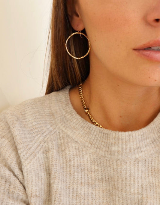 A close-up view of a woman's face features a large, textured gold hoop earring and a gold chain necklace, set against a soft, light-colored sweater. Her hair partially frames her face.