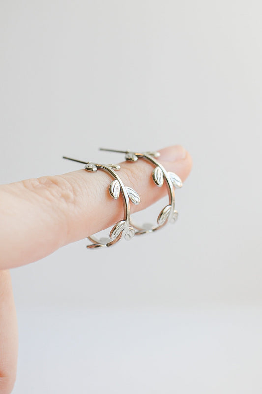 A hand holds a pair of silver hoop earrings designed with leaf-like embellishments, showcasing their delicate style against a softly blurred, light background.