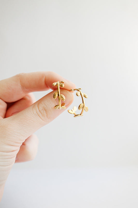 A hand holds a pair of delicate, gold hoop earrings designed like intertwining vines with small leaves, showcased against a light, neutral background.