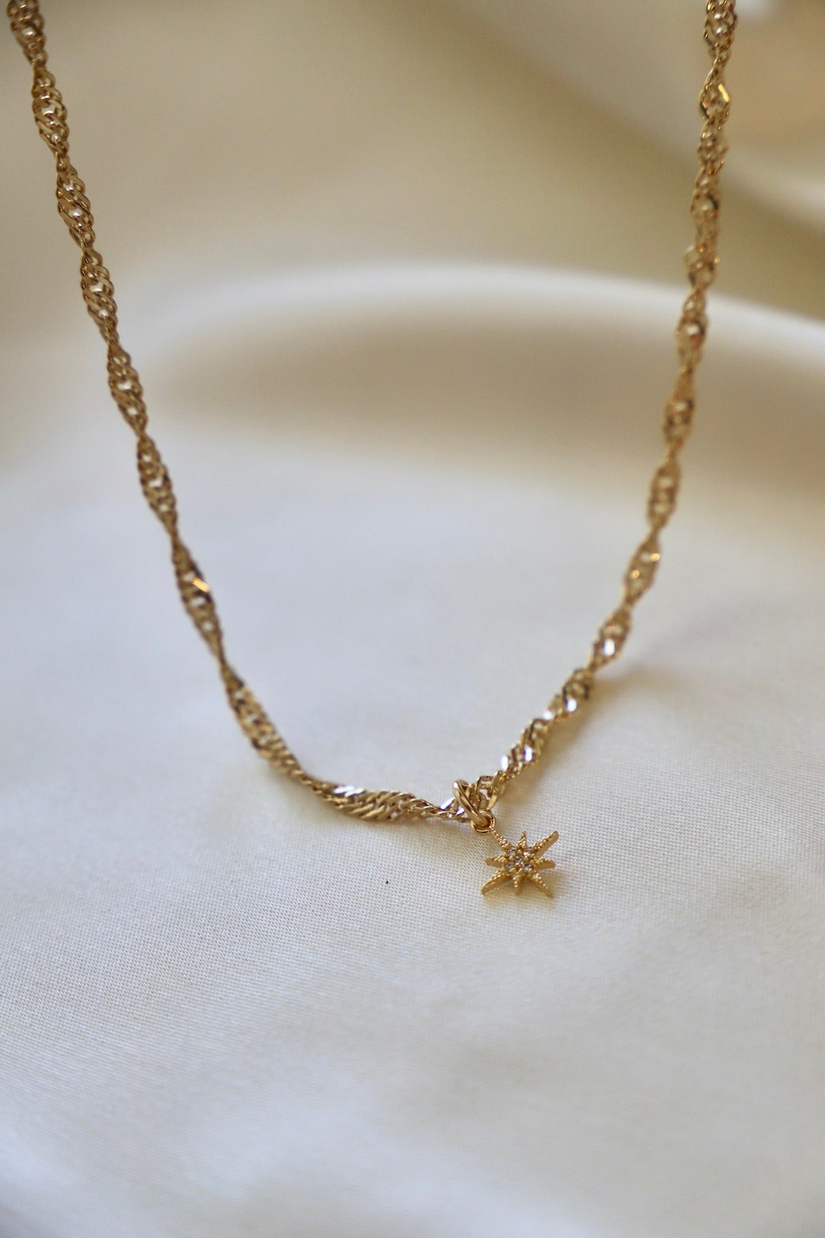 A twisted gold necklace features a star-shaped pendant, resting on a soft, light-colored fabric backdrop, highlighting its intricate design and shiny surface.