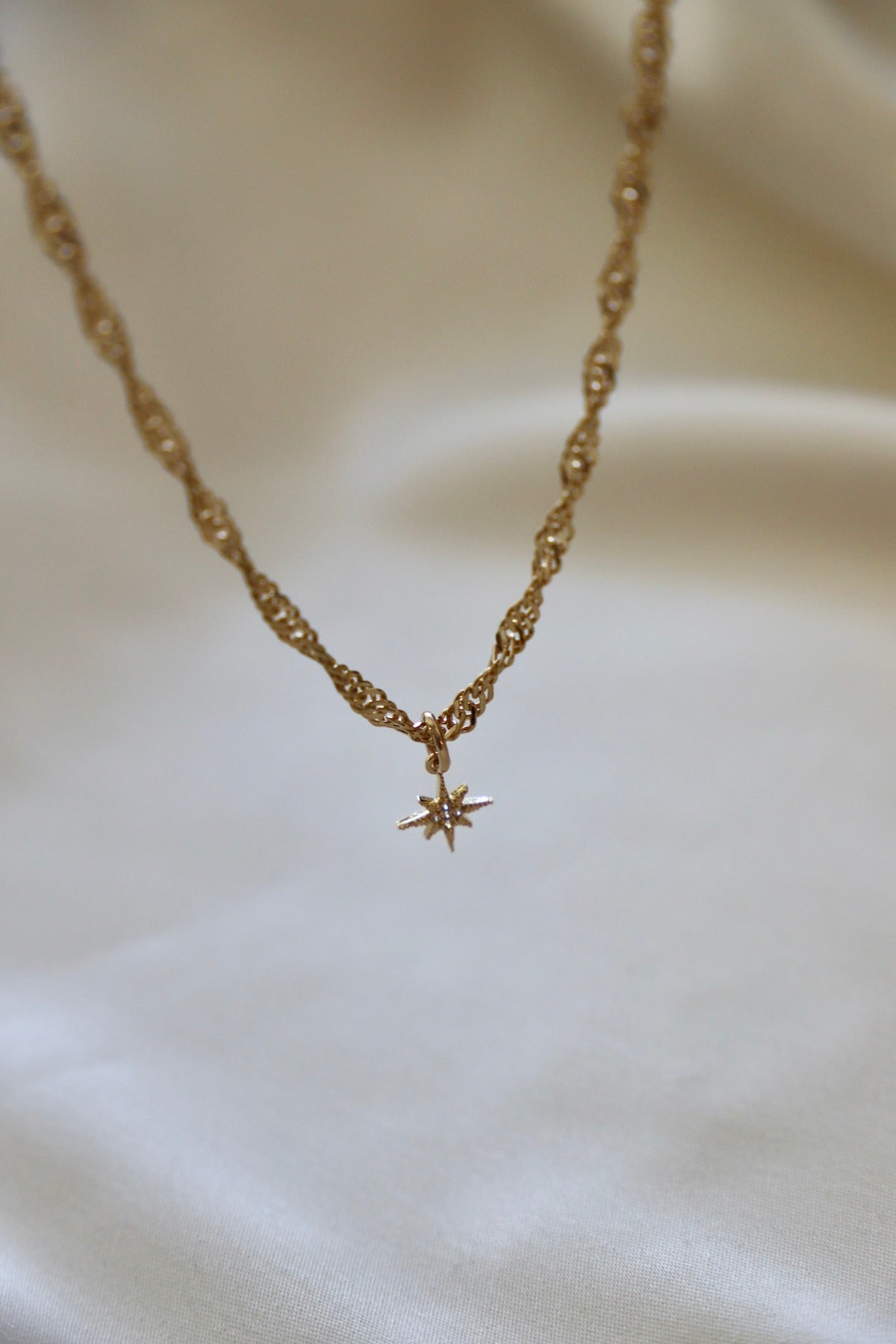 A twisted gold necklace features a small star-shaped pendant, resting on a soft, light-colored fabric background, creating an elegant and delicate appearance.