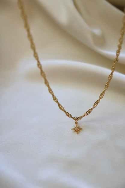 A gold necklace with a twisted chain design hangs elegantly, featuring a small star-shaped pendant. It rests on a smooth, light-colored fabric in a soft, serene setting.