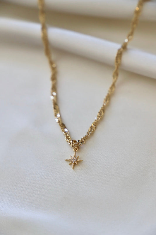 A gold necklace with a twisted chain features a star-shaped pendant with sparkling details, resting on a smooth, light-colored fabric background.