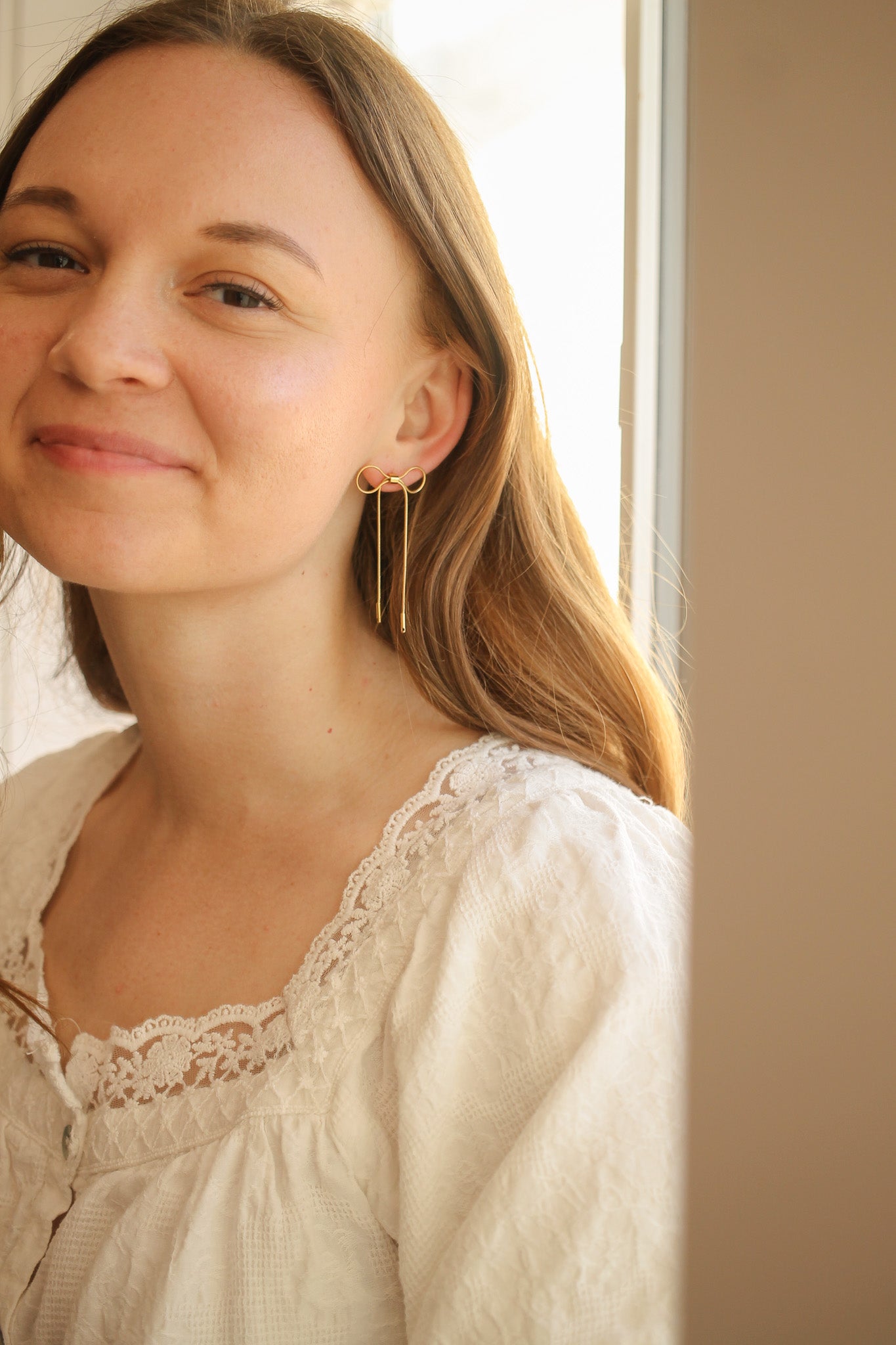 A woman with long hair smiles, wearing a unique gold earring shaped like an infinity symbol with hanging strands. She is positioned near a window, highlighting natural light.