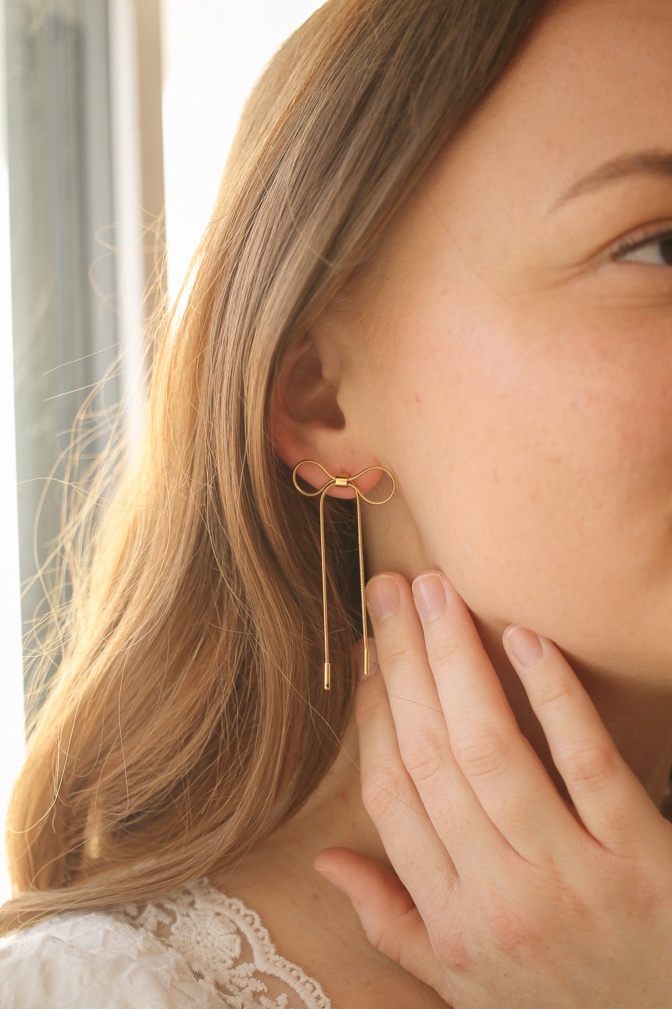 A woman gently touches her cheek with one hand, showcasing a pair of elegant gold bow-shaped earrings that hang just below her earlobes, against a softly lit background.