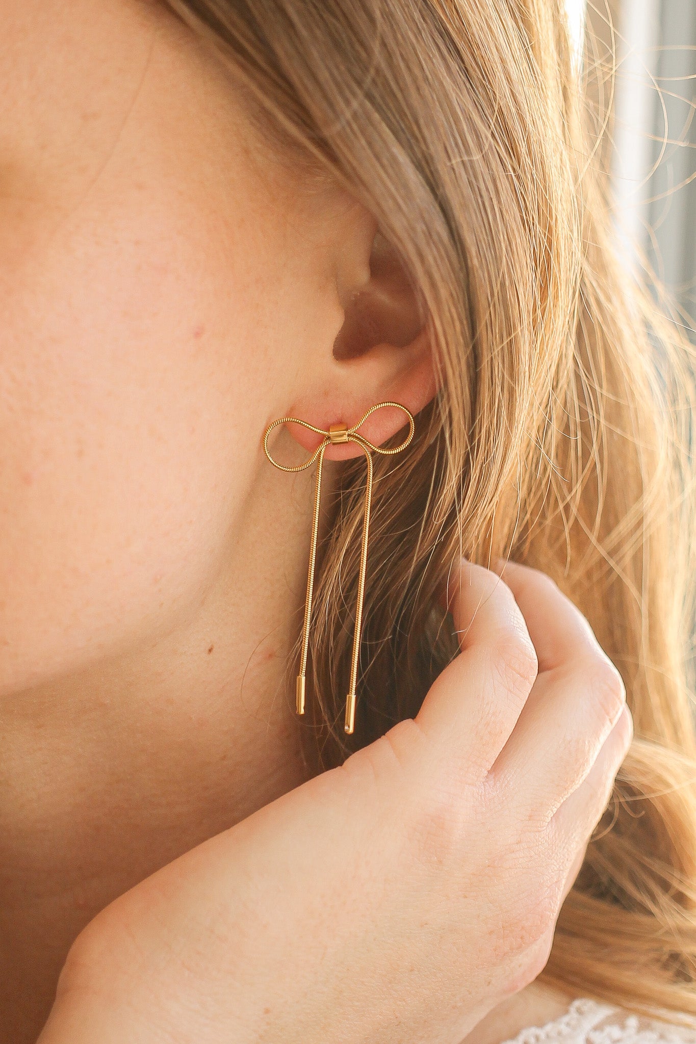 A pair of gold earrings shaped like a bow dangles from the wearer’s ear. The person gently touches the earring while sunlight reflects on their hair, creating a soft ambiance.