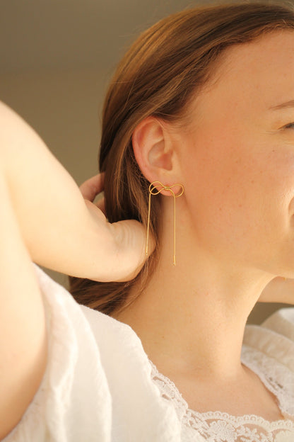 A woman is adjusting her hair while wearing a gold infinity-shaped earring, which features a long dangling piece. The background is softly blurred, enhancing her serene expression.