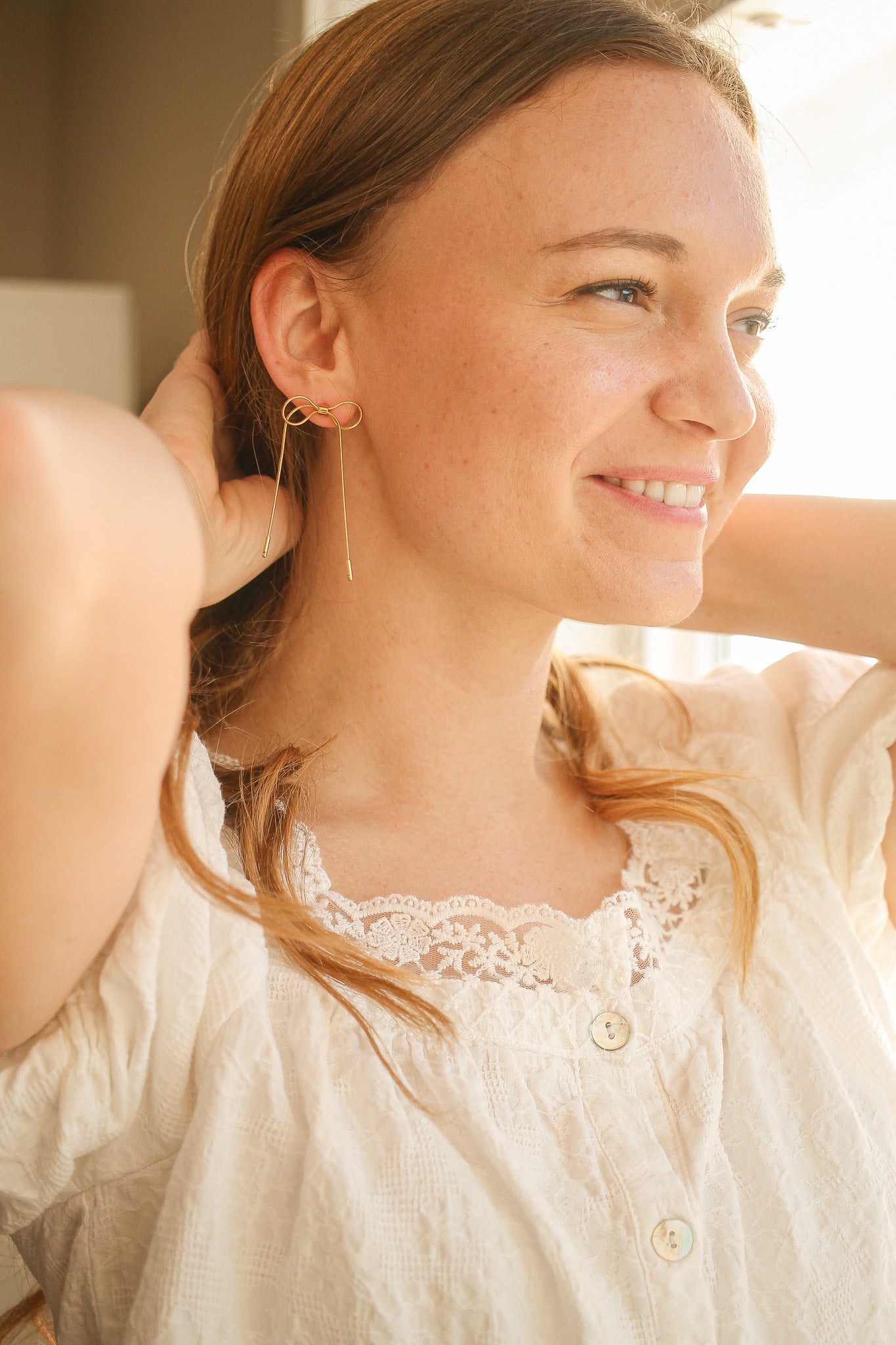 A woman with long hair is smiling while adjusting her hair. She wears unique gold earrings shaped like an infinity symbol. The setting is bright, suggesting a comfortable indoor space.