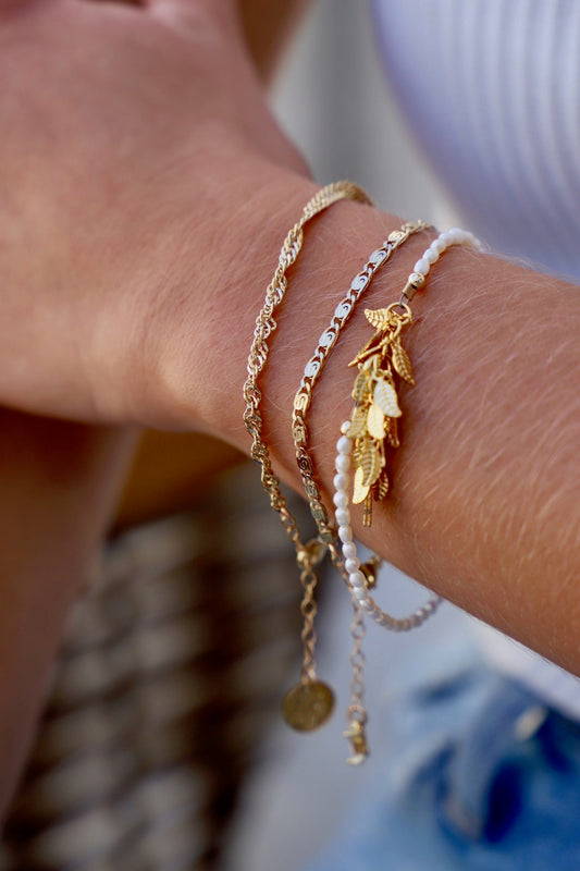 A wrist adorned with multiple delicate bracelets, including chains and a charm featuring leaf designs, rests against a blurred background of soft colors and patterns.