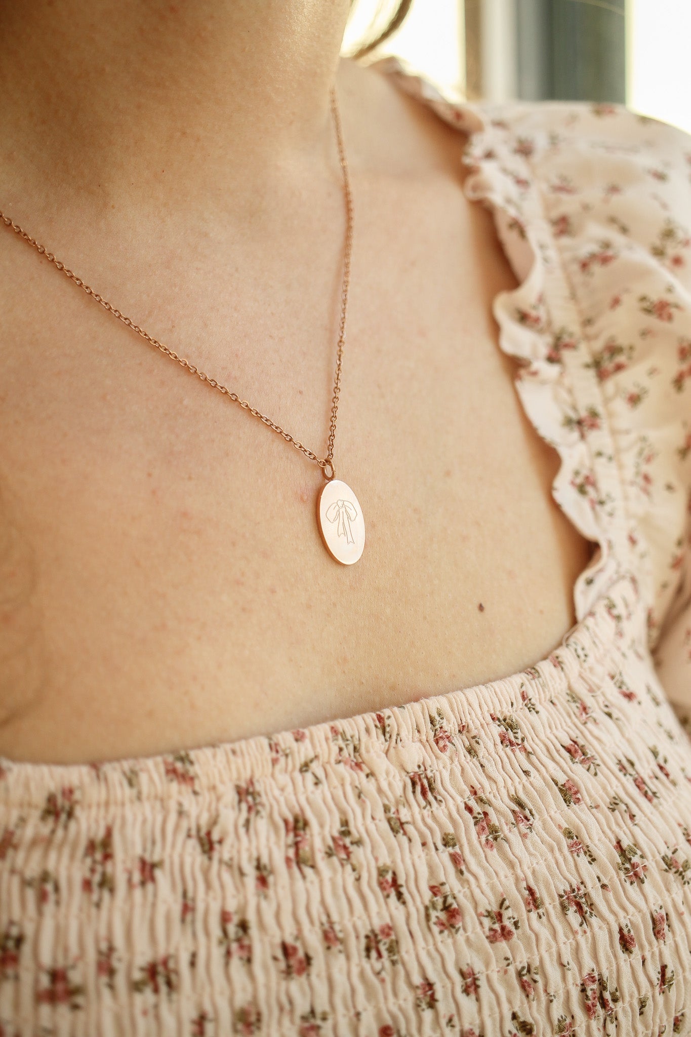 A delicate necklace with an oval pendant featuring a floral design hangs around a person's neck, adorned by a soft, floral-patterned blouse in a bright, well-lit environment.