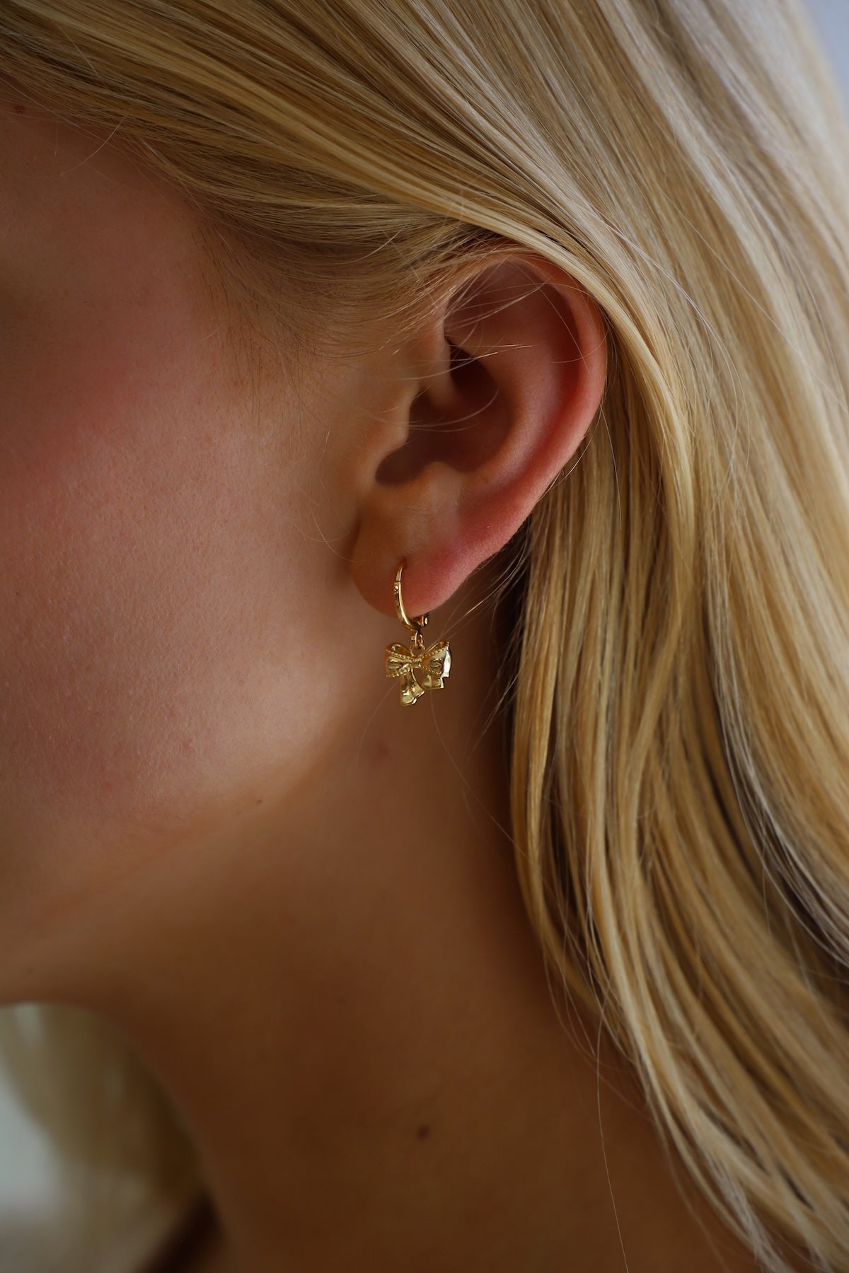 A close-up of a womanâ€™s ear adorned with a delicate gold hoop earring featuring a small butterfly charm, set against a softly blurred background of light colors.