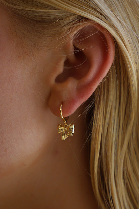 A golden bow-shaped earring hangs from a person's earlobe, highlighting delicate features against a backdrop of straight, blonde hair cascading down.