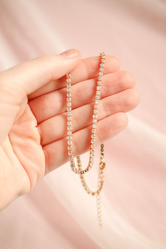 A hand holds a sparkling, gold-accented chain with shimmering stones, showcasing its elegant design against a soft pink background. The chain drapes delicately between the fingers.