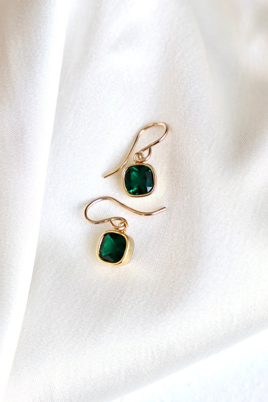 Two elegant gold earrings featuring green square-shaped stones are resting on a soft, white satin fabric, reflecting light and adding a touch of luxury to the scene.