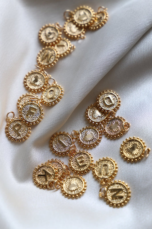 Gold circular pendants, each featuring an embossed letter, are scattered across a smooth, light-colored fabric. The pendants, intricately designed with beaded edges, reflect light softly.