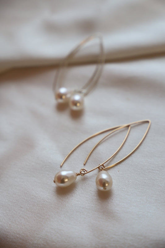 Golden pearl earrings rest on a soft, light-colored fabric. One pair features two drop-shaped pearls, while another has a simple hoop design, creating an elegant, understated arrangement.