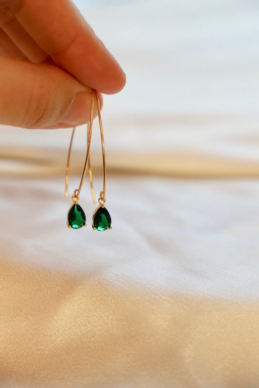 A hand holds a pair of elegant green gemstone earrings. The earrings feature teardrop-shaped stones suspended from thin gold hooks, set against a soft, silky beige surface.