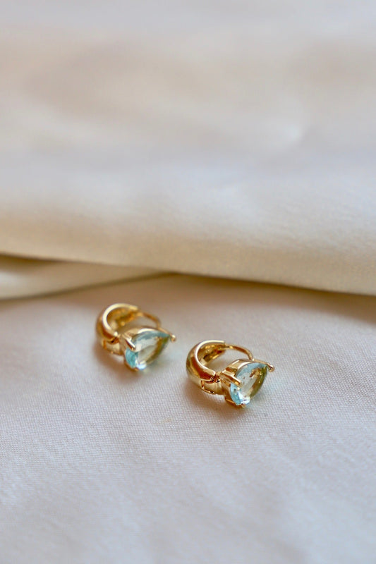 Two gold hoop earrings with pear-shaped light blue gemstones are positioned on a soft, cream-colored fabric, suggesting elegance and luxury in a delicate, inviting setting.