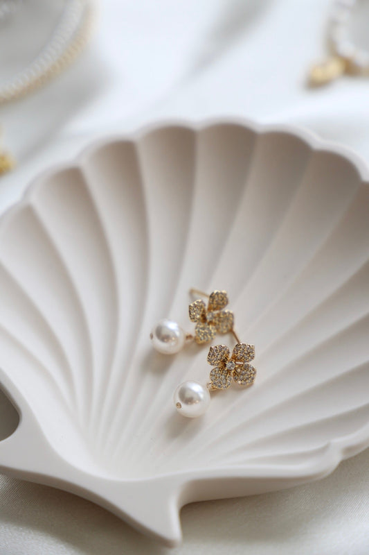 Two elegant earrings, one featuring a pearl and the other a sparkly flower design, rest delicately on a white, shell-shaped dish, set against a soft, light-colored background.