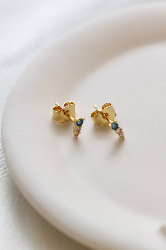 A pair of gold earrings rests on a light-colored dish. They feature a unique design with a small blue stone and decorative details, showcasing elegance against a soft fabric backdrop.