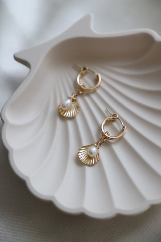 Two gold earrings featuring shell designs and pearl accents rest in a white shell-shaped dish on a soft fabric surface, showcasing delicate craftsmanship and elegance.