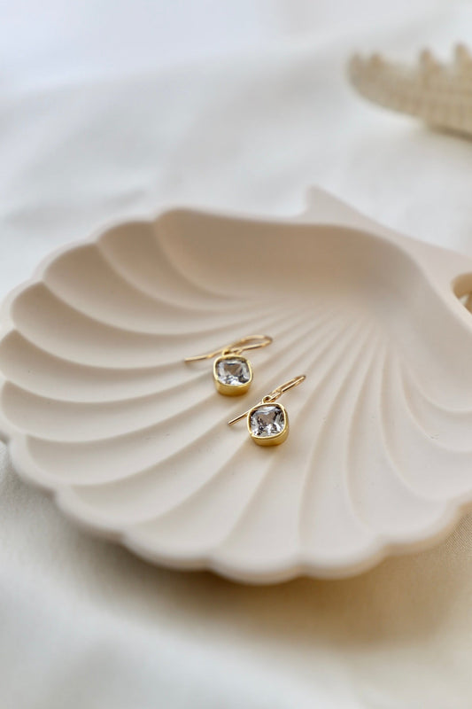 Gold earrings with square gemstones rest on a textured, clam-shaped dish. The dish sits on a soft, neutral fabric surface, creating an elegant and serene display.