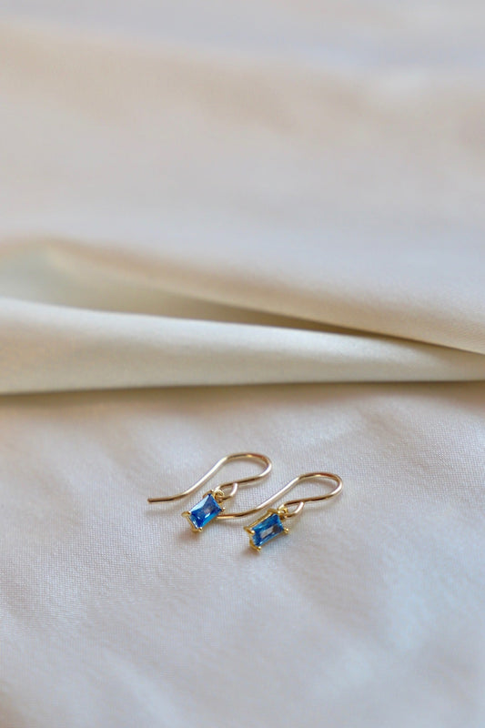 Two gold earrings with rectangular blue gemstones rest on a smooth, light-colored fabric. The scene conveys elegance and simplicity, highlighting the jewelry's color against the soft texture.