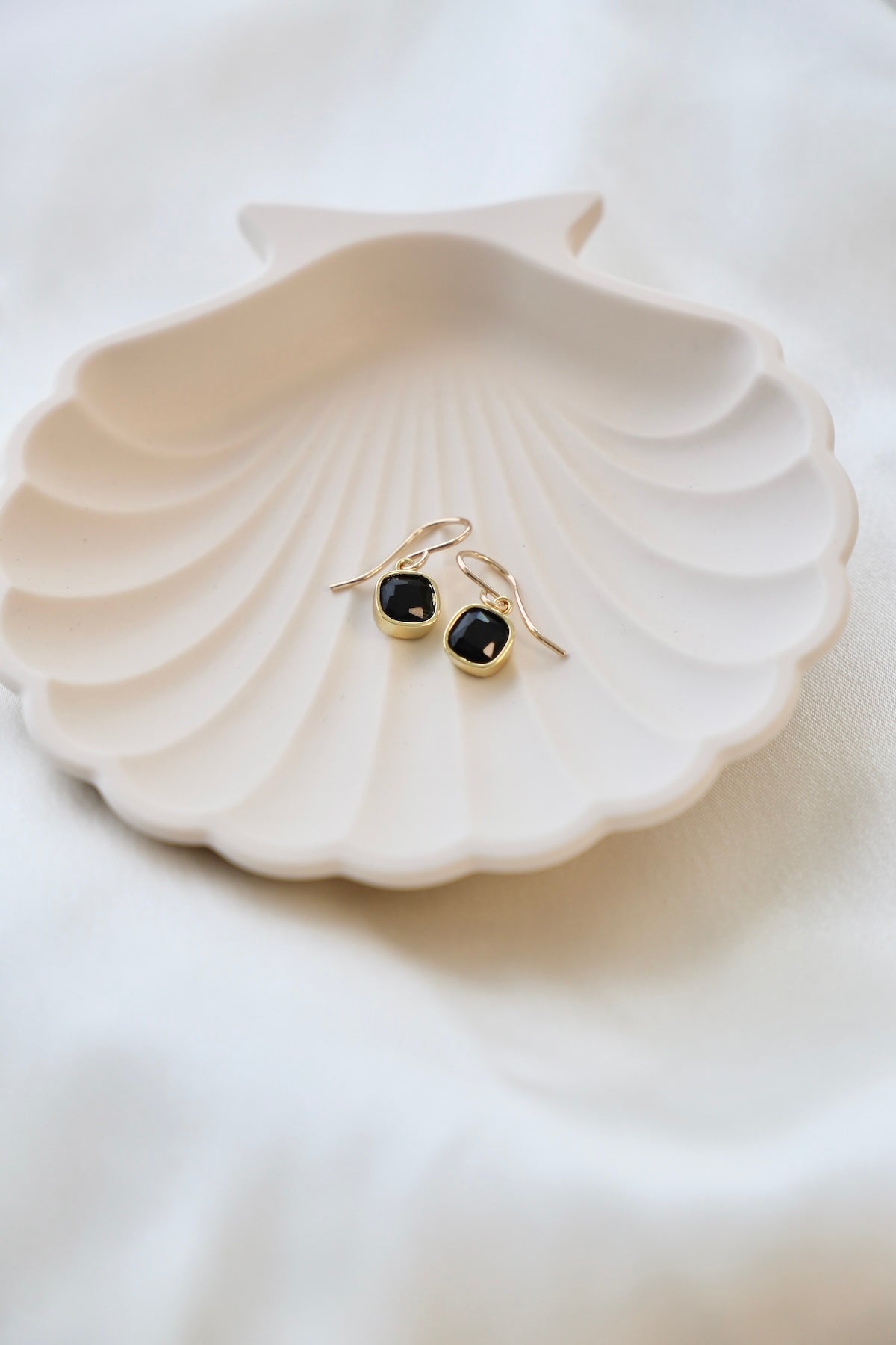 Two elegant black earrings with gold accents rest on a scalloped white dish. The dish is placed on a soft, light-colored fabric, creating a serene and minimalist arrangement.