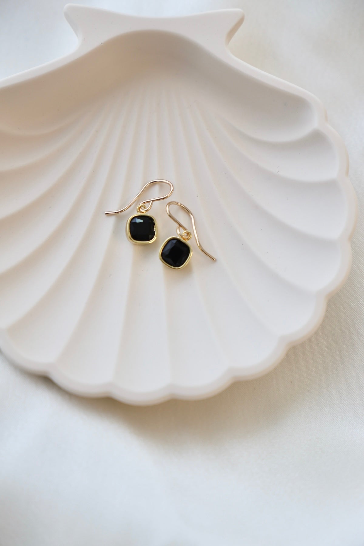 A delicate, white shell-shaped dish holds a pair of gold and black gemstone earrings. The soft, light background adds an elegant touch to the display.