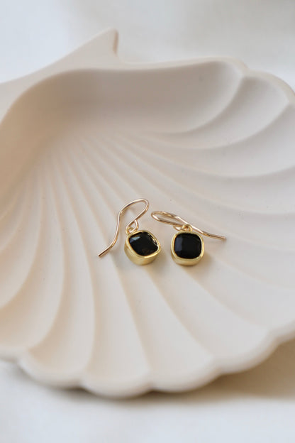 Two elegant black stone earrings with gold settings rest on a textured, beige shell-shaped dish, creating a soft, sophisticated display against a neutral background.