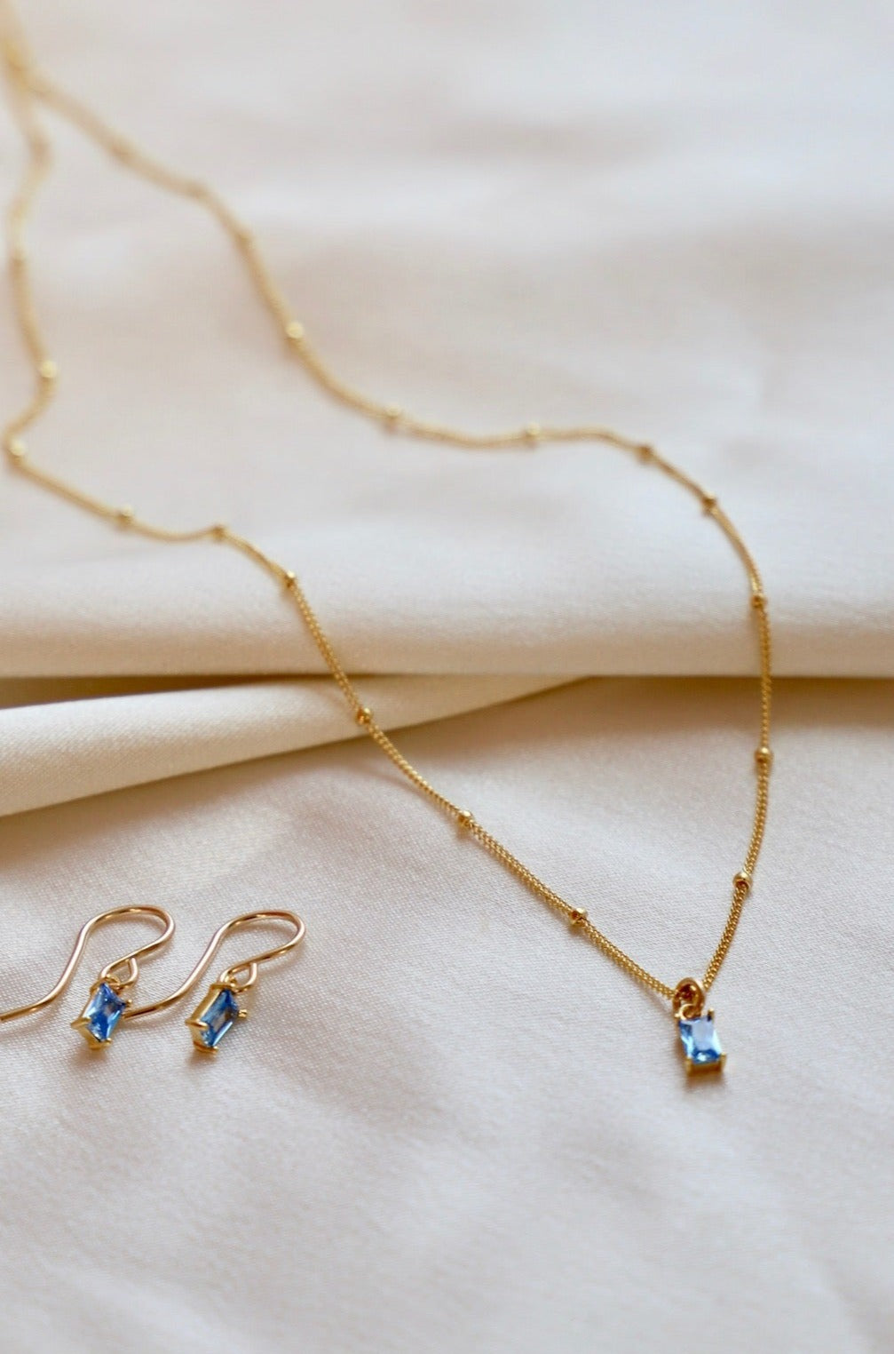 A gold necklace and matching earrings are elegantly displayed on soft, cream-colored fabric. The jewelry features delicate blue gemstones, showcasing a luxurious yet minimalistic design.