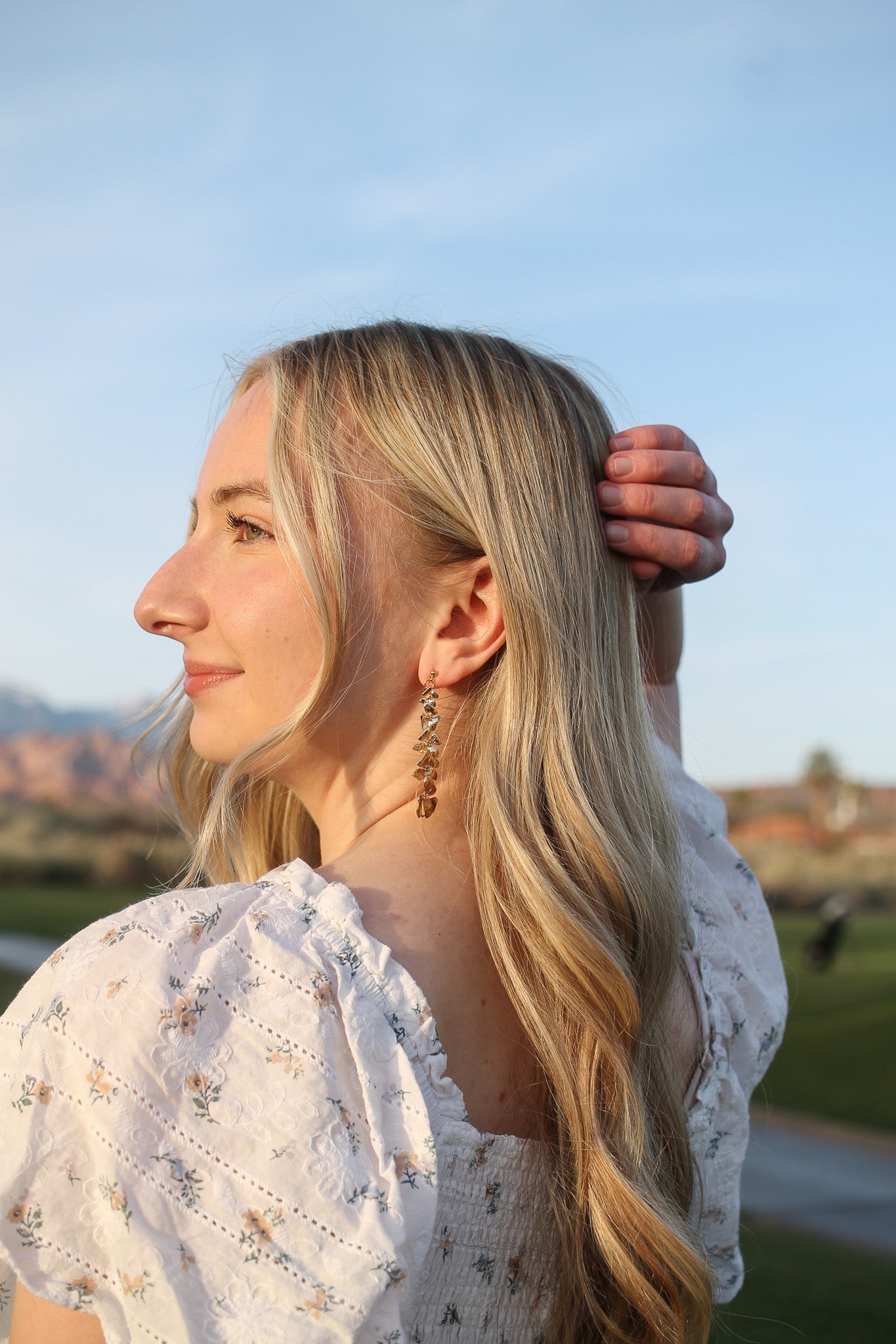 A woman with long, wavy blonde hair, wearing decorative earrings, gently brushes her hair behind her ear. She stands outdoors, with a clear blue sky and green grass in the background.