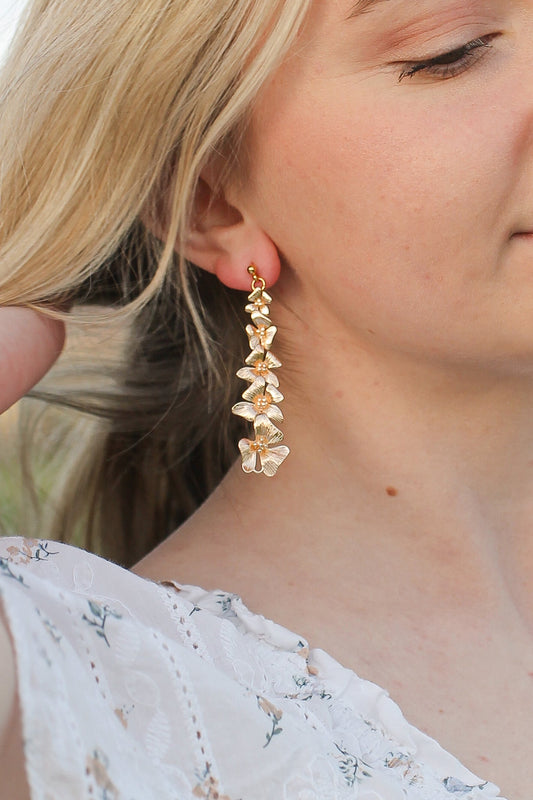 A gold flower-shaped earring hangs from the ear of a woman. She lightly touches her hair, set against a softly blurred natural background.