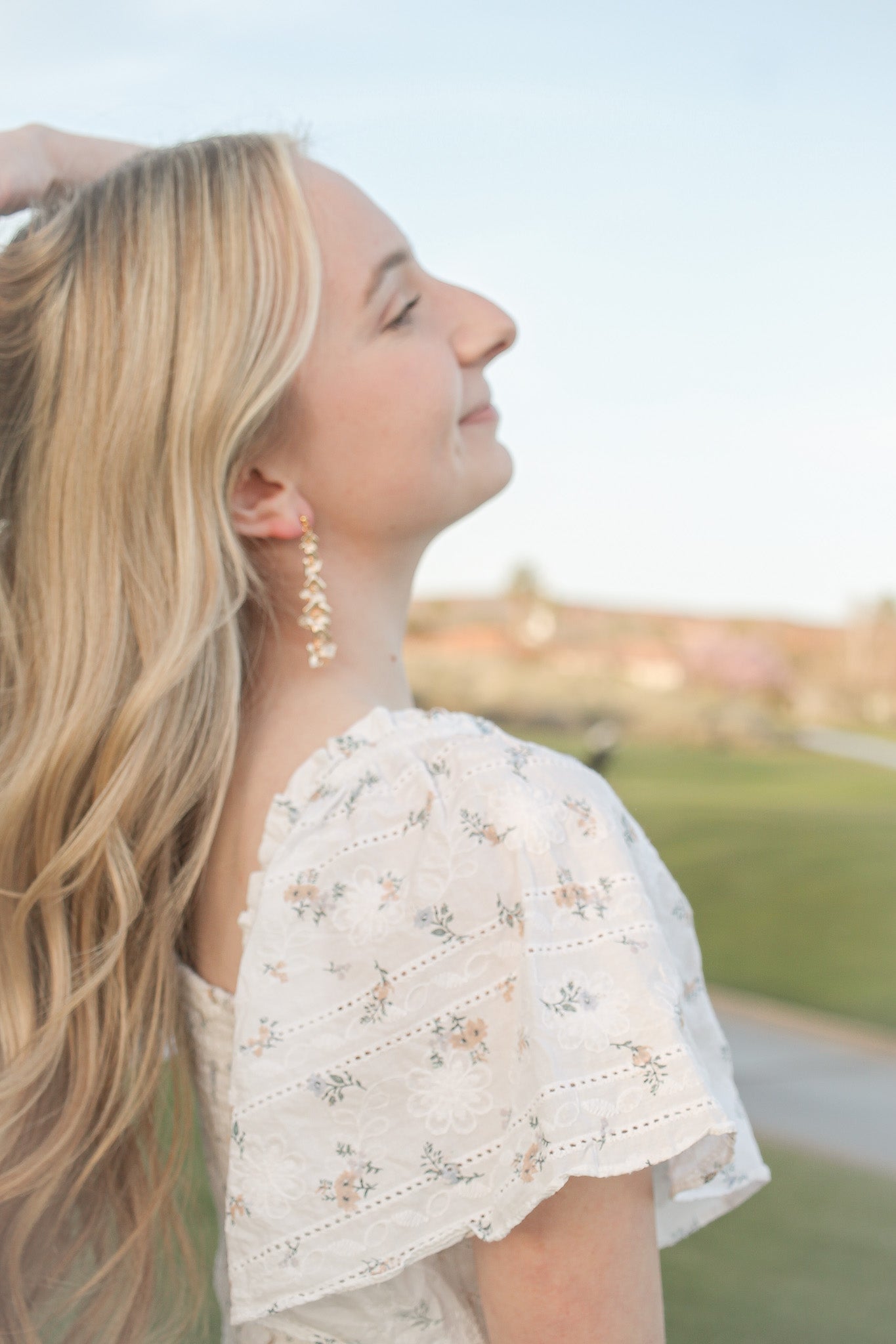 A woman with long, wavy hair and floral-patterned attire stands outside, gently touching her hair, smiling serenely against a blurred green landscape and soft sky.