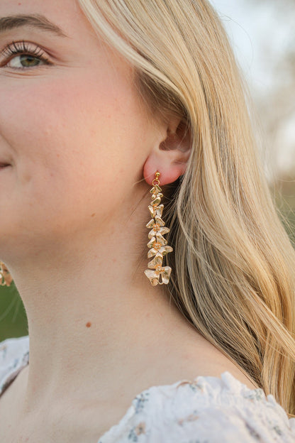 Long, golden floral earrings dangle from a smiling woman’s ear as her light blonde hair cascades down her shoulder, set against a blurred outdoor background.