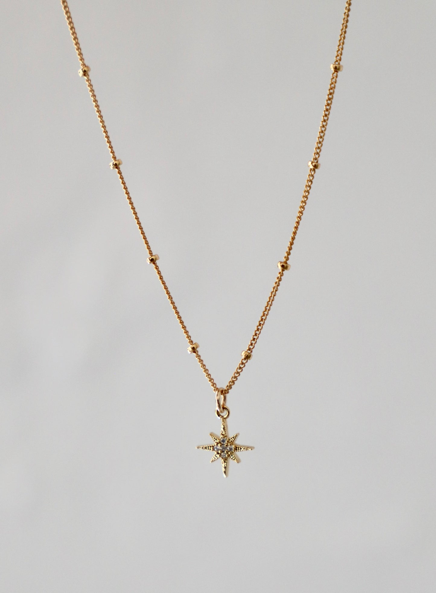 A delicate gold necklace features a star-shaped pendant adorned with small stones, suspended from a finely linked chain. The background is light and neutral, emphasizing the jewelry's shine.