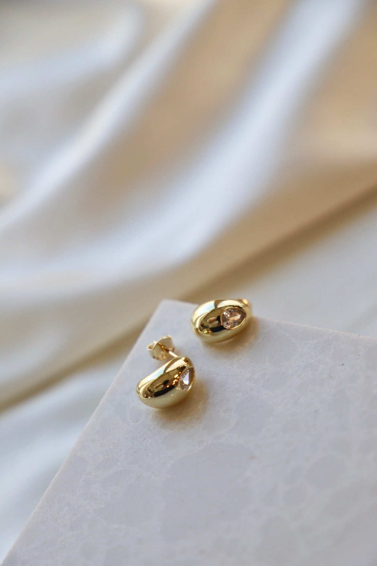 Gold earrings with a teardrop shape and a central stone rest on a textured white surface, surrounded by soft, flowing fabric in shades of cream and beige.