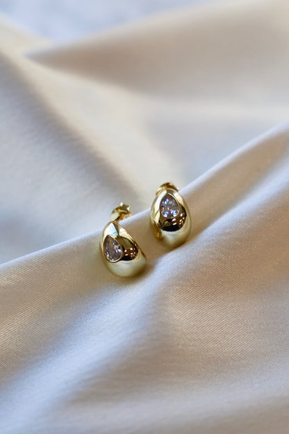 Two golden hoop earrings, featuring a teardrop shape with a clear gemstone, rest elegantly on smooth, shimmering fabric, reflecting soft light in a subtle, sophisticated display.