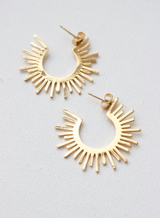 Gold hoop earrings are displayed, featuring a sunburst design with protruding rays. They are positioned on a textured white surface, emphasizing their shine and unique shape.