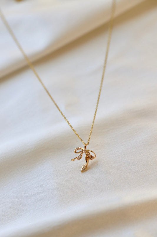 A delicate gold necklace with a flower-shaped pendant rests on a pale, textured surface. The pendant features intricate details and small sparkling accents, reflecting light softly.