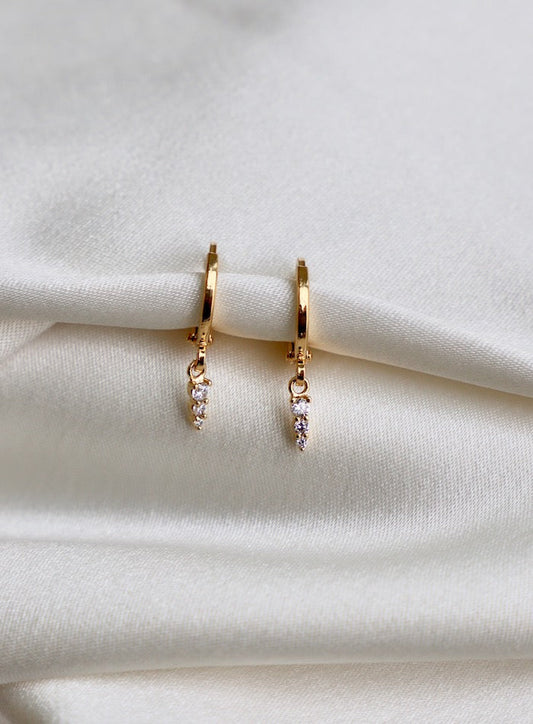 Gold hoop earrings with small, pointed, sparkling charms hang delicately on a smooth, white satin fabric, creating a simple yet elegant display against the soft background.