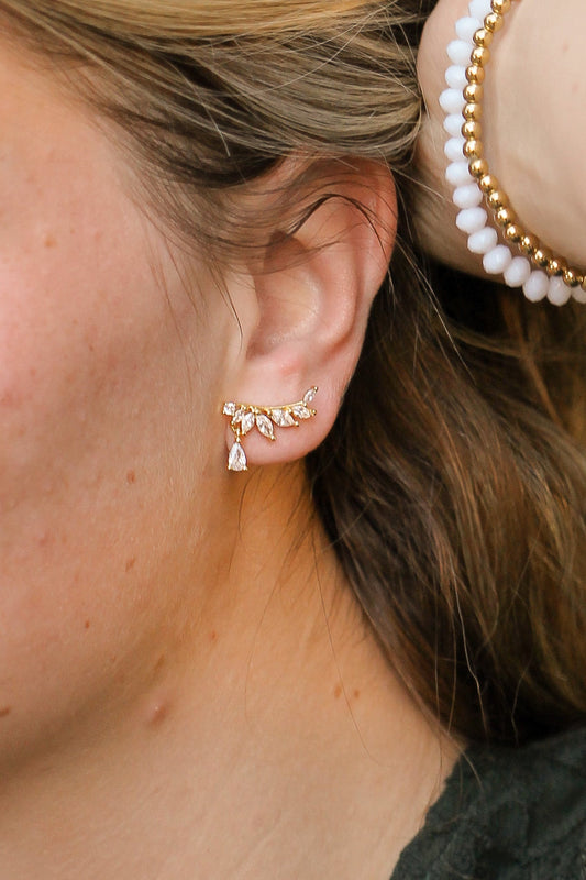 Gold earrings shaped like a branch adorned with sparkling stones are worn on a person’s ear, with a light brown hair strand visible, set against a neutral background.