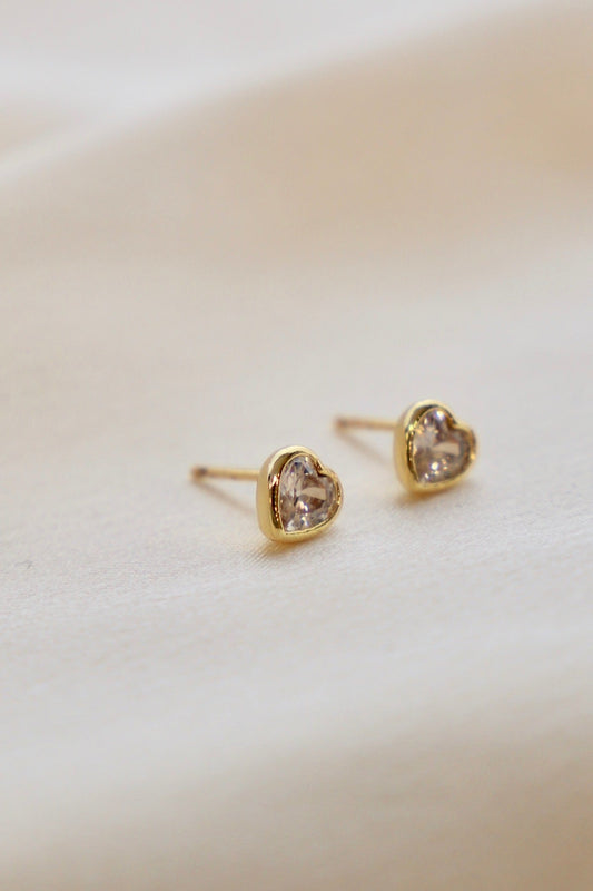 Two heart-shaped gold earrings with clear gems sit elegantly on a smooth, light-colored fabric, showcasing their shine and intricate design in a soft, neutral setting.