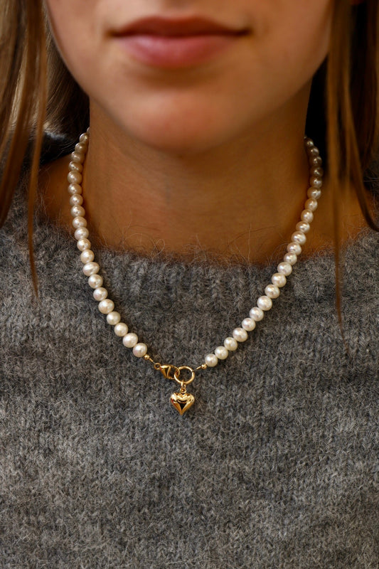 A pearl necklace adorned with a heart-shaped charm rests on a person's neck, complementing a grey, knitted sweater. The background is softly blurred, focusing on the necklace and the skin tone.