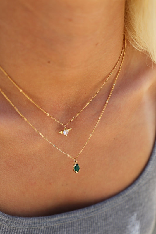 Gold necklaces featuring a small bird pendant with a gemstone and an oval green gemstone pendant hang above a gray tank top, set against warm skin tones.