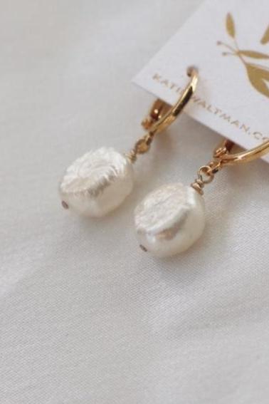 Two earrings, featuring round, iridescent pearls, hang from gold clasps. They rest on a soft, light fabric background and are attached to a card with a minimalist design.