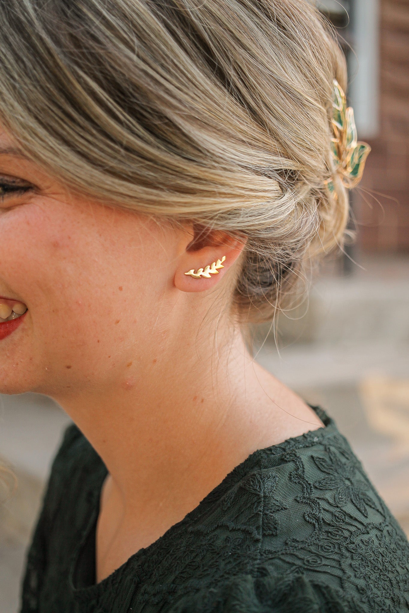 A woman with light blonde hair styled in a bun smiles, wearing a delicate gold leaf earring. She is dressed in a textured dark green top, with a blurred outdoor setting behind her.